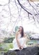 A woman in a white dress sitting on a rock under a tree.